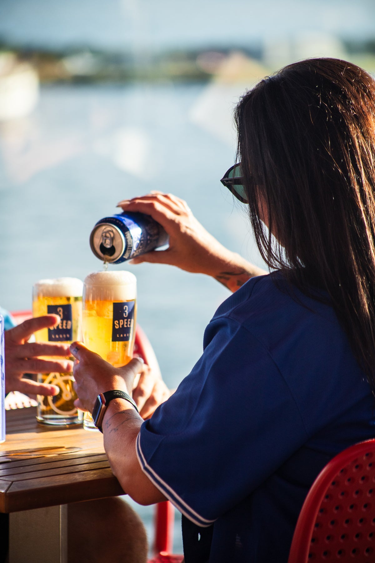 Person sitting at a table by a body of water, pouring a beer from a can.