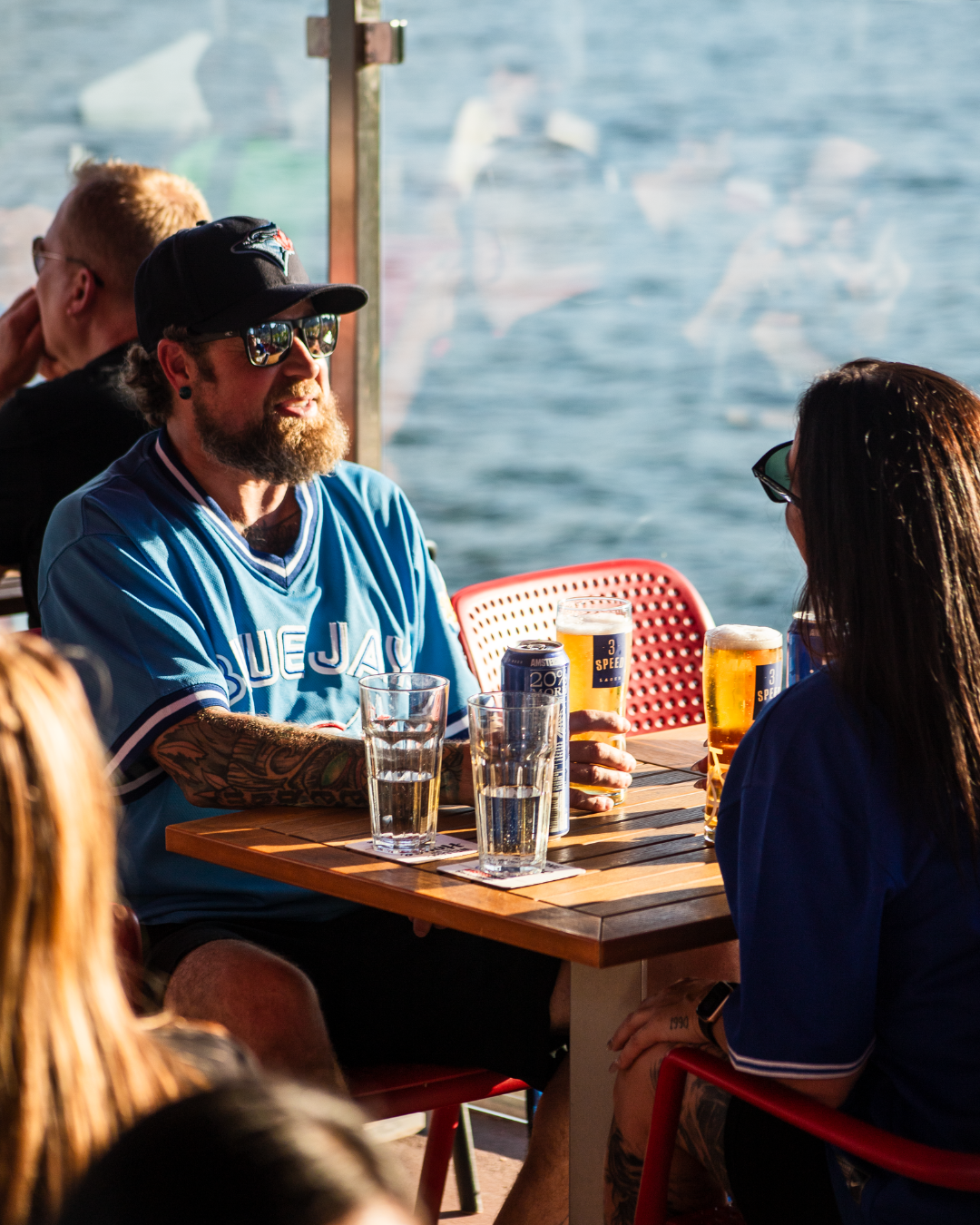 People sitting at a table by the water with 3 Speed beers while wearing a Blue Jays jersey