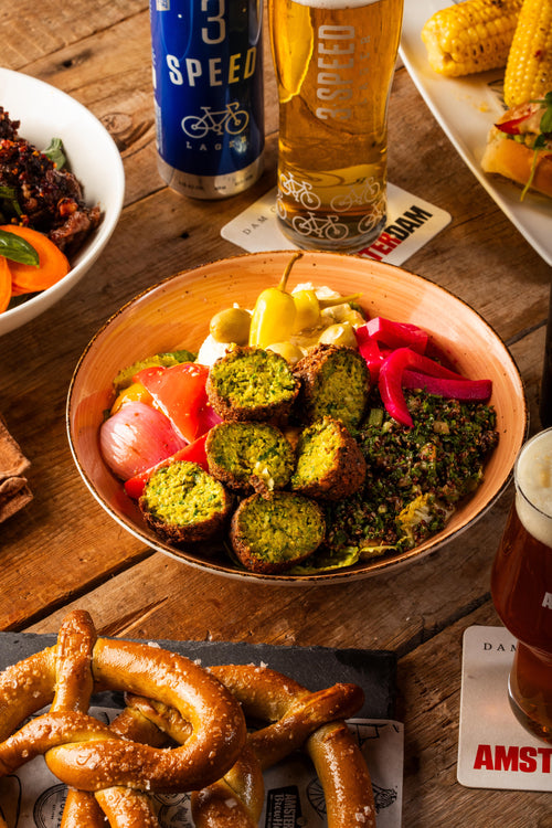 A bowl of falafel, tomatoes, olives, and greens with pretzels on a table next to Amsterdam's 3 Speed beer. 